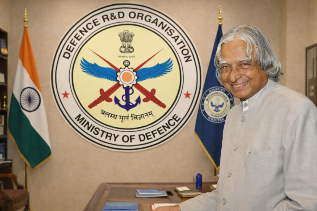 An elderly Indian scientist and leader standing in an office with the Defence Research and Development Organisation emblem and Indian national flag in the background.