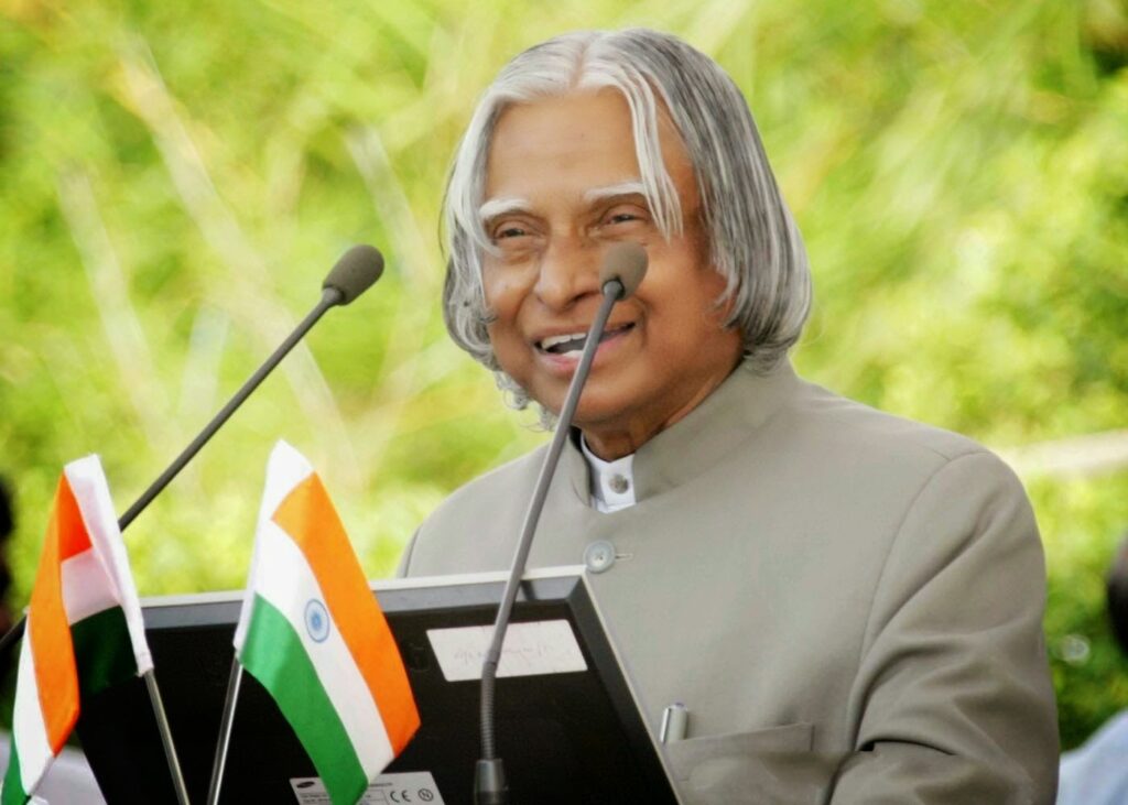 An elderly Indian statesman smiling while delivering a speech at a podium, with Indian national flags placed in front.